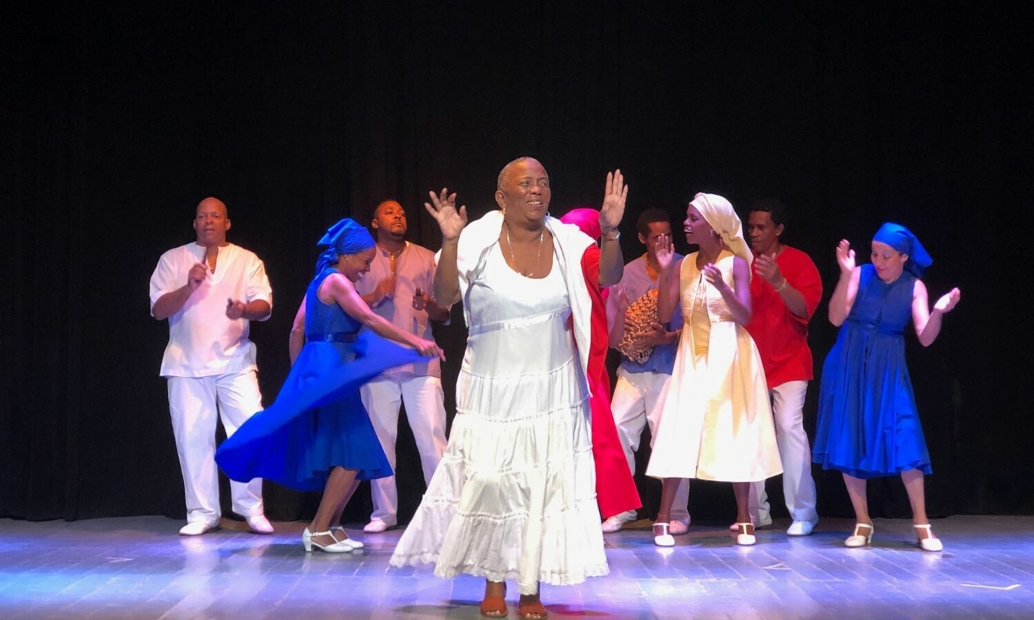 A woman in a white dress with short hair dancing with an ensemble of other women and men behind her.