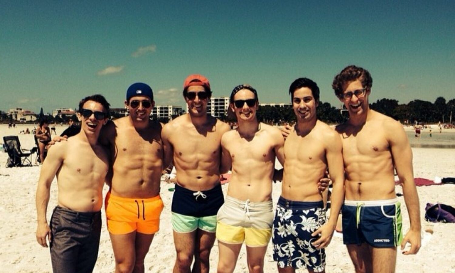 Six shirtless men in swim trunks at the beach.