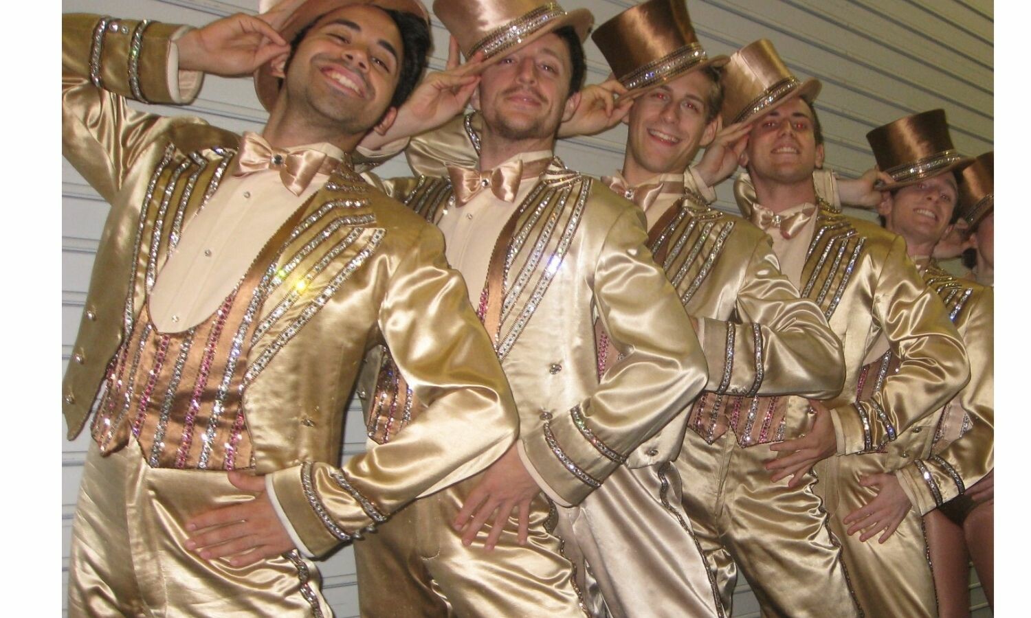 Five men in a line, each wearing a golden three-piece suit and tipping his top hat to the side. 
