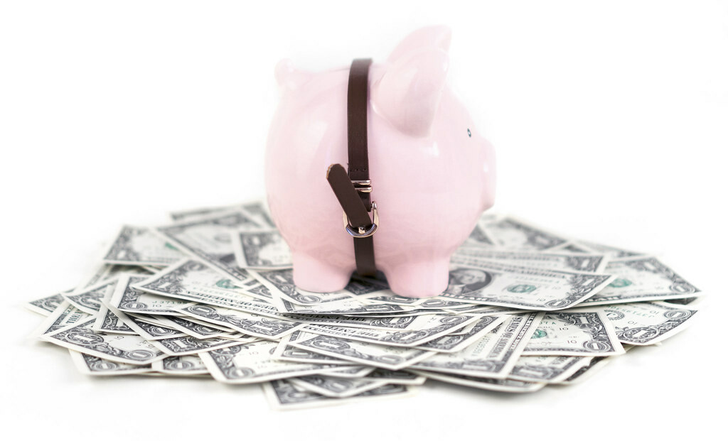 A pink piggy bank with a black belt stands on top of a pile of one dollar bills.