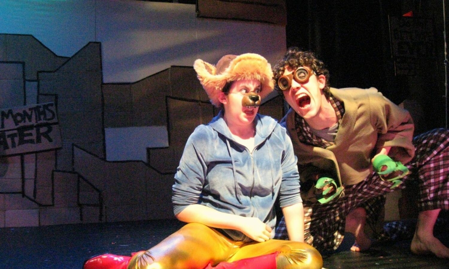 An actor dressed as a lion sitting onstage next to another actor dressed in brown. Both appear to be screaming.