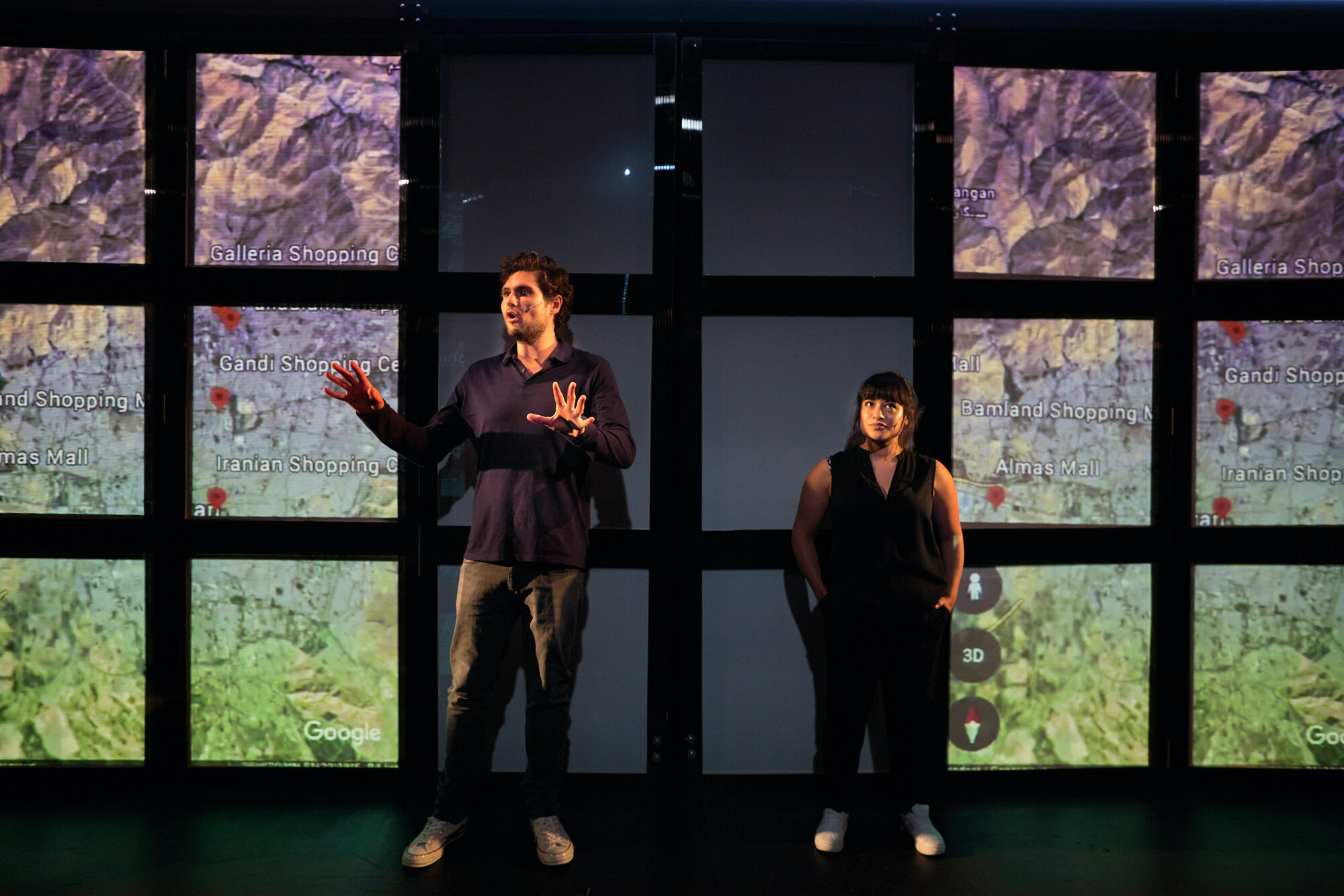 A man (left) and woman (right) standing in front of several screens, some with parts of a map on display.