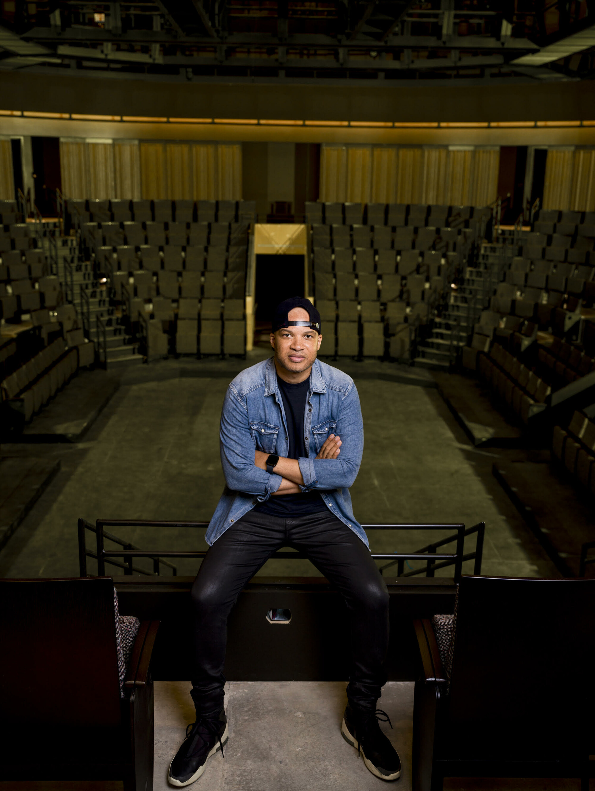 Individual wearing blue jean jacket, black baseball cap and, black shirt and pants sitting on railing in theatre.