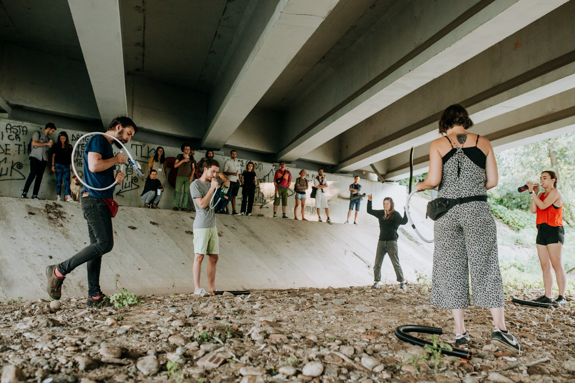 Five people stand in a circle under a bridge with an audience standing above them.