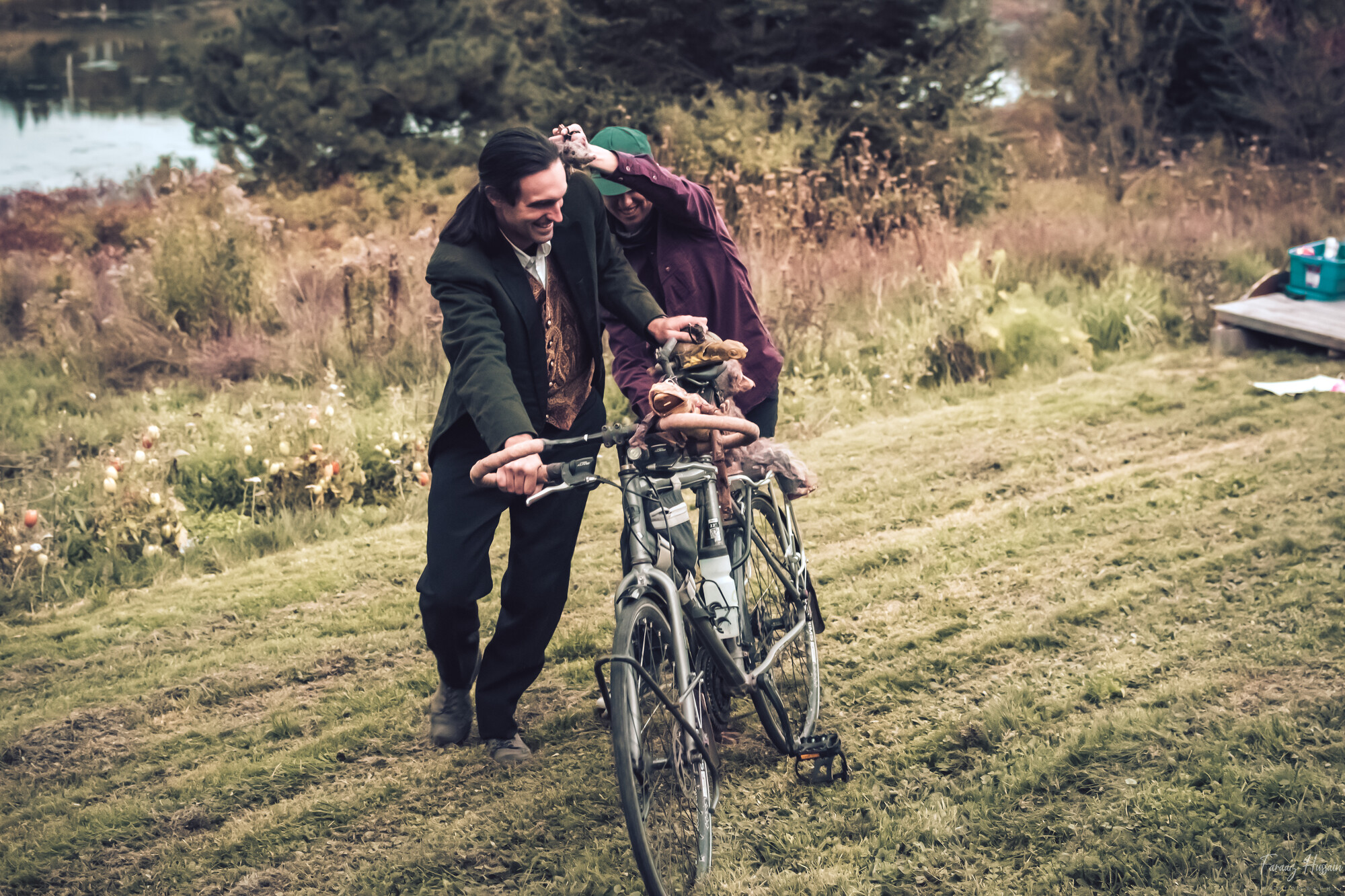 Two men pushing a bicycle through a field.
