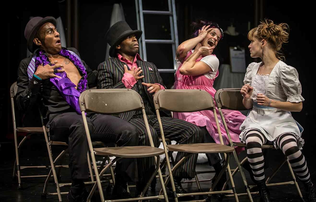 Four actors sitting in folding chairs performing on stage.