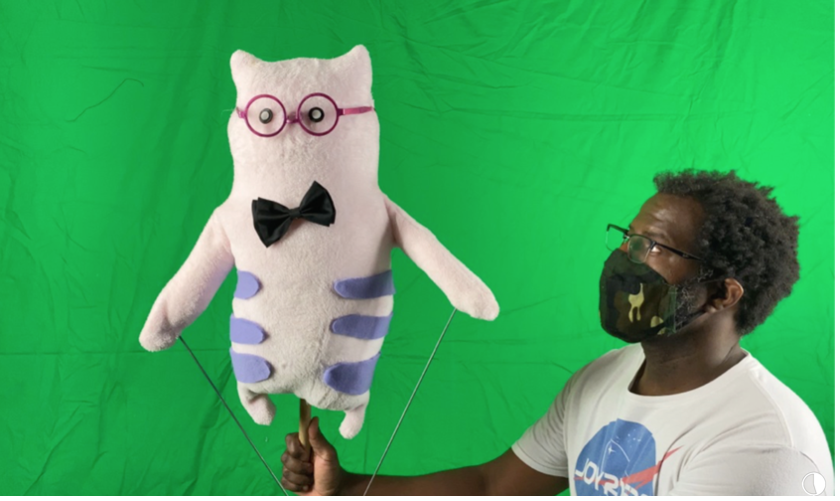 A man holds a puppet above his head in front of a green screen.