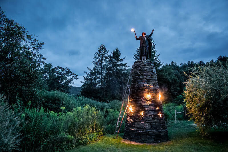 A man carrying a torch stands on top of a small stone tower.