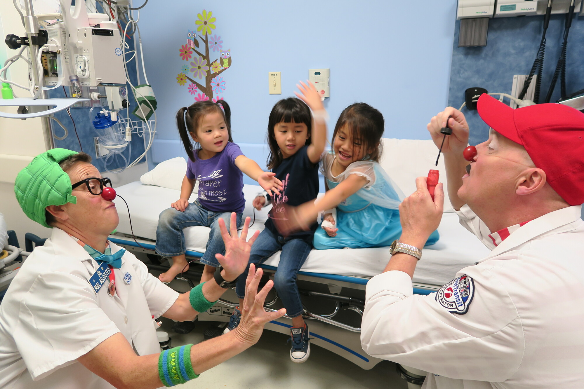 Two clowns entertaining three children in a hospital room.