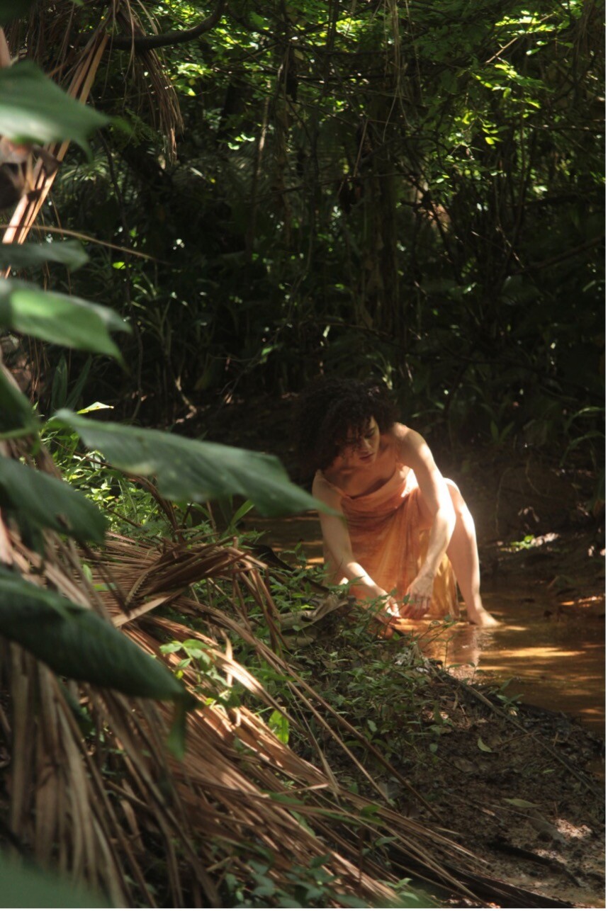 A woman bends down in a river surrounded by trees.