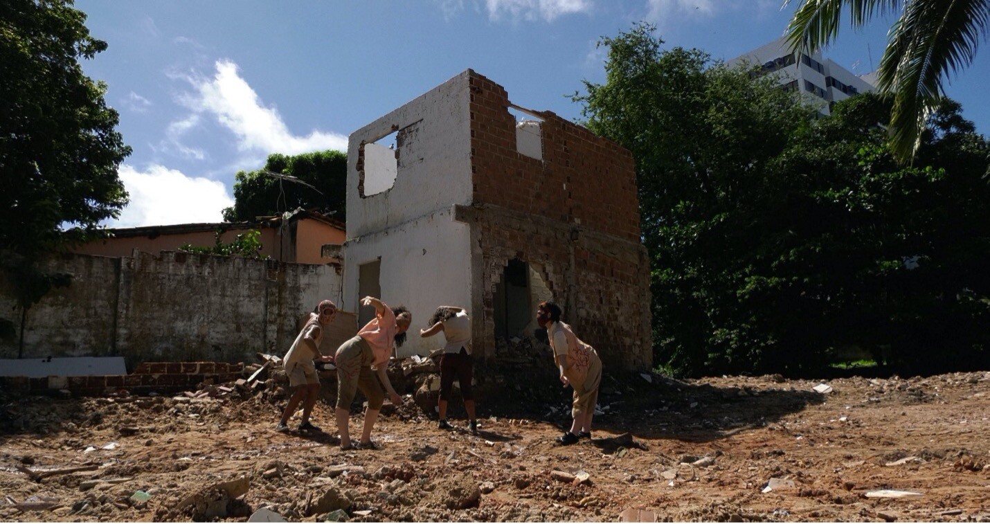 Four people posing or dancing outside of a damaged building.