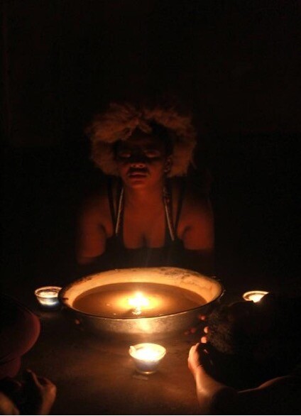 A woman sits in the dark in front of a candlelit bowl of water.