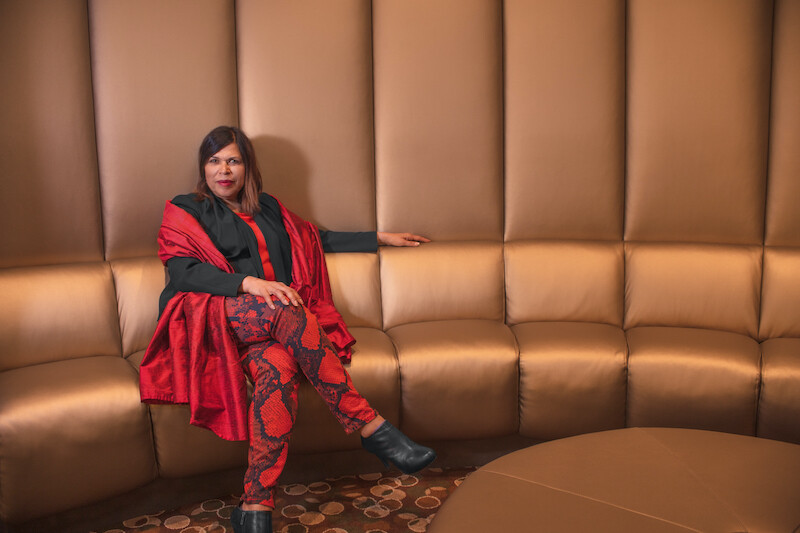 Diane Rodriguez sits on a tan couch wearing red pants, black long sleeve shirt, and red shawl.