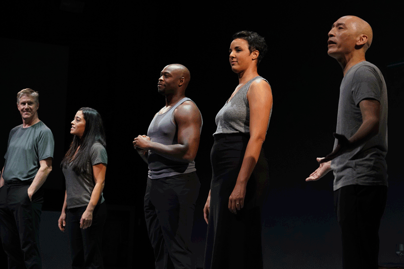 Five actors in black clothes stand on a dark stage.