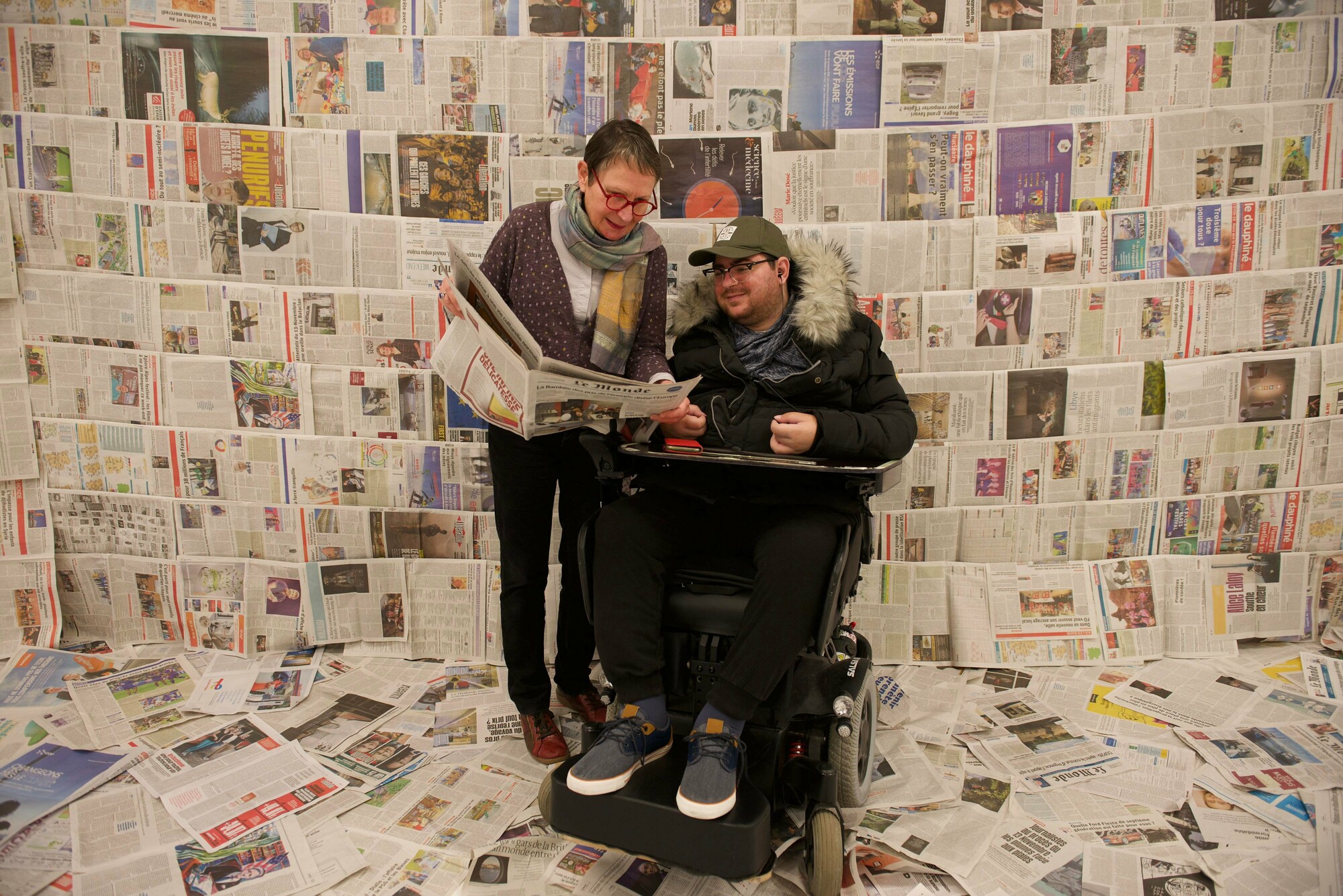 Two people standing in a room with newspaper on the walls.
