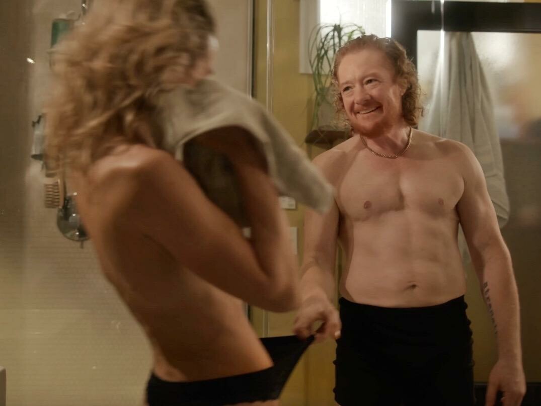 Two topless actors smiling at one another.
