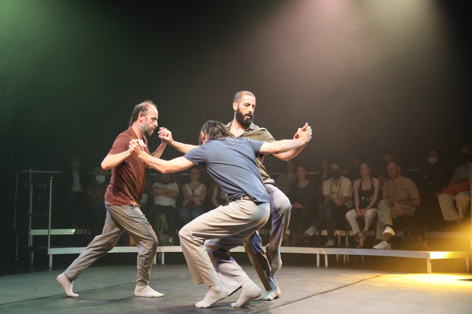Three bearded men dance around each other in a circle under a spotlight onstage.