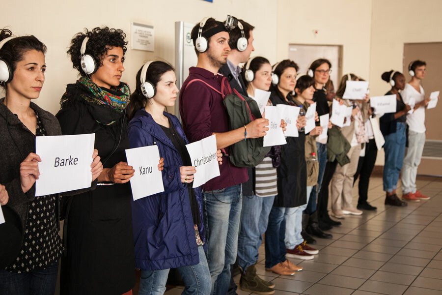 Fourteen people wearing headphones stand in a line holding sheets of paper with different words on them.