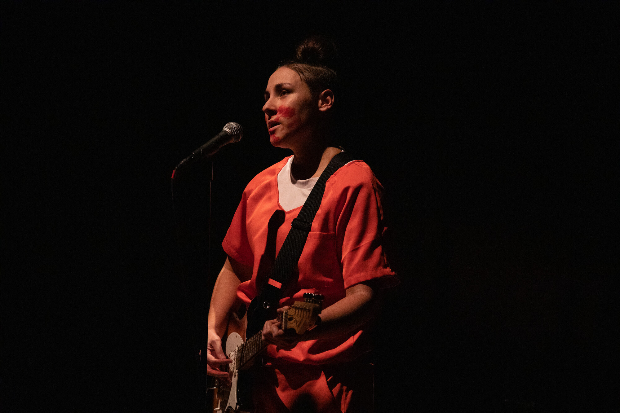 A woman in a long orange shirt playing the guitar under a spotlight.