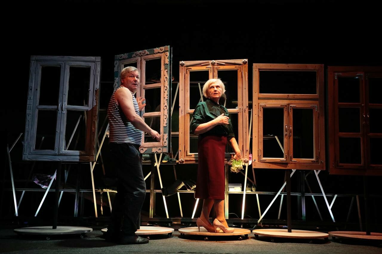 One performer speaks over her shoulder to another in front of a series of window frames on stage.