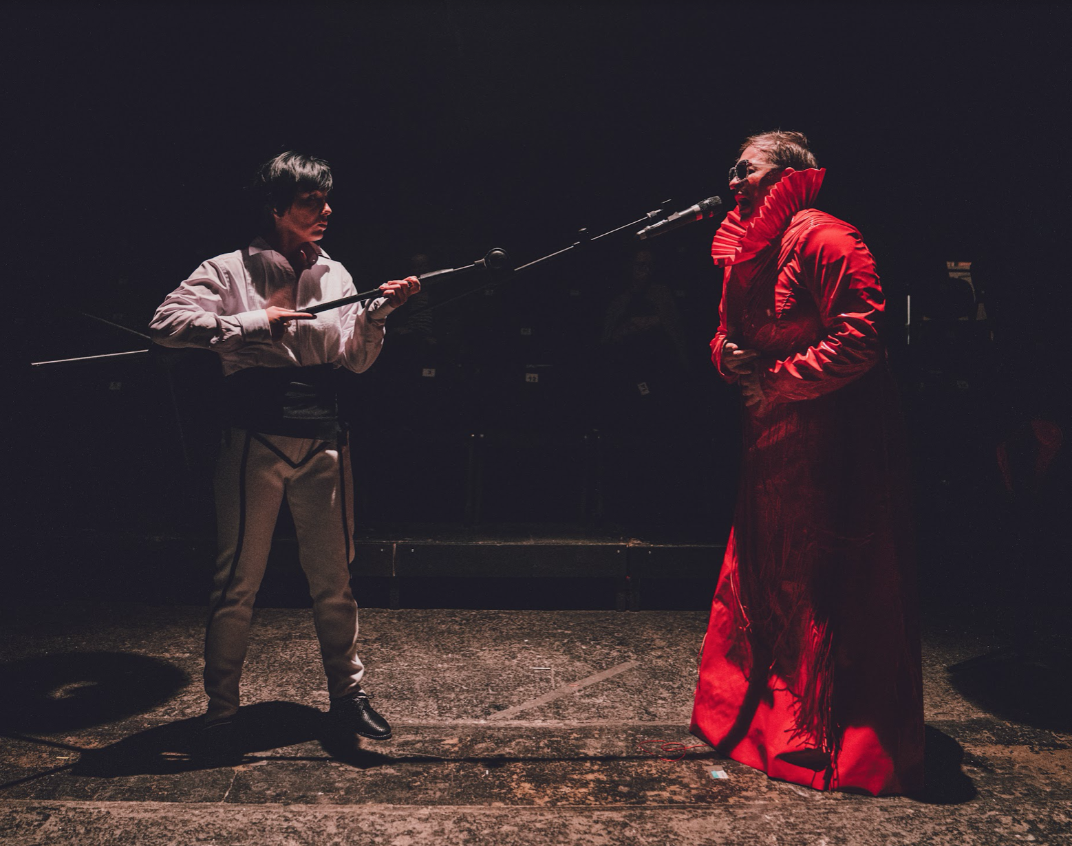 An actor dressed in white points a microphone on a long stick at another actor dressed in red.