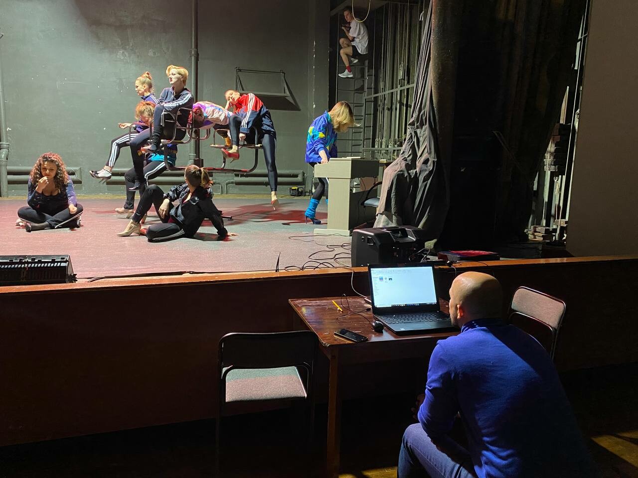 A person sits in front of a laptop watching actors rehearse on stage.