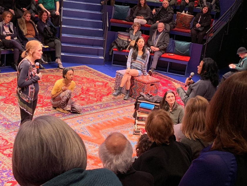 Five women gather on a carpeted stage as the audience watches.