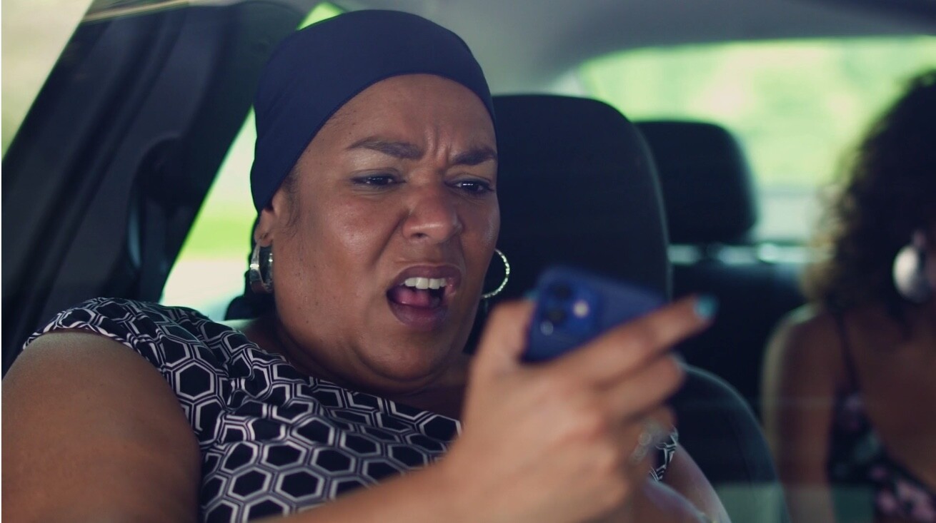 A woman seated in a car looks at a phone screen, shocked.