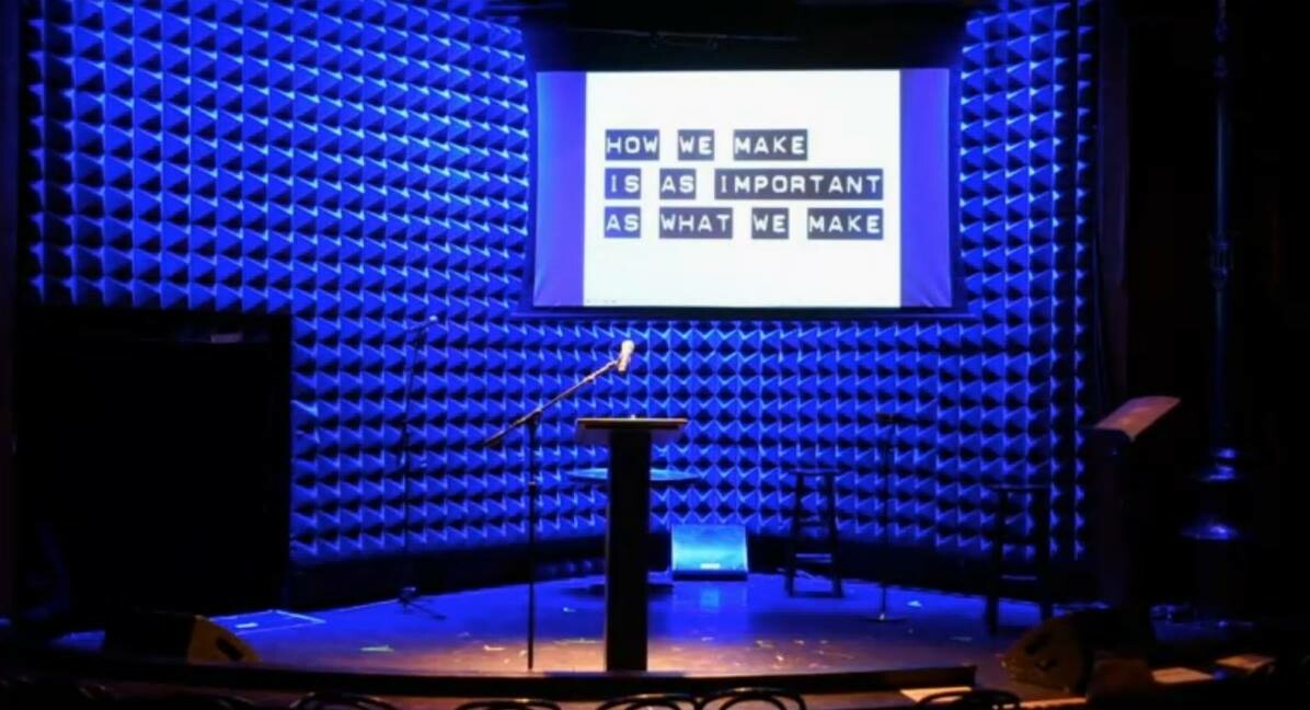 Podium on a stage with a screen behind that reads "How we make is as important as what we make."