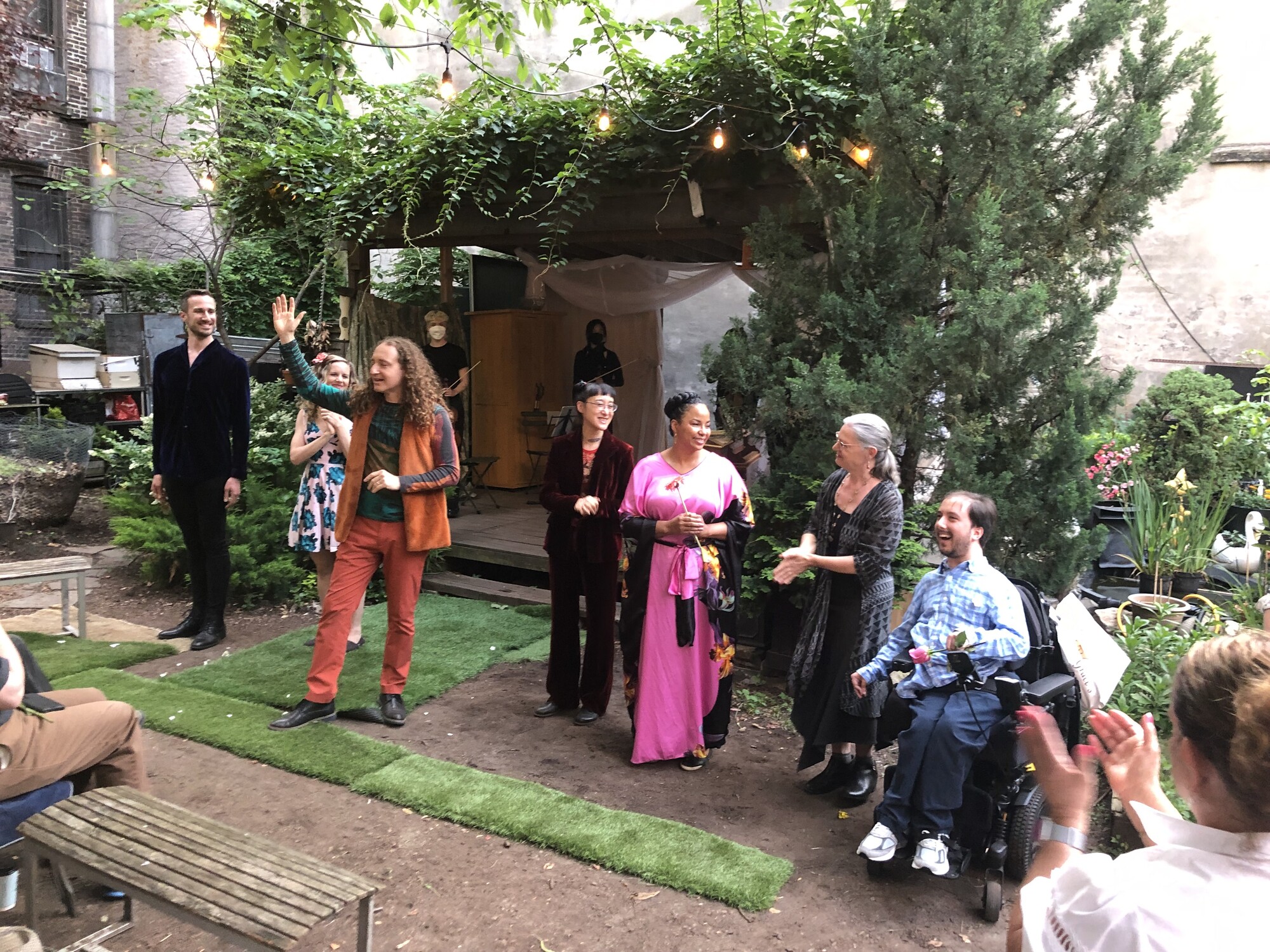 A wide shot of a verdant outdoor stage at Campos Community Garden as the setting for a baroque opera - the performers are taking their bows at the end surrounded by flowers and lush bushes that frame a wooden stage, upon which stands a baroque string orchestra.