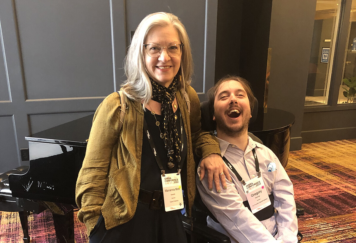 A light skinned woman with long silver hair and a black dress stands proudly with a light skinned man in a blue shirt, sitting in a wheelchair. His arms are to one side in a gesture of a hug while she has her arm over him and his chair, leaning in. They are both smiling and proud about the opera they have just produced and are about to present to the public.