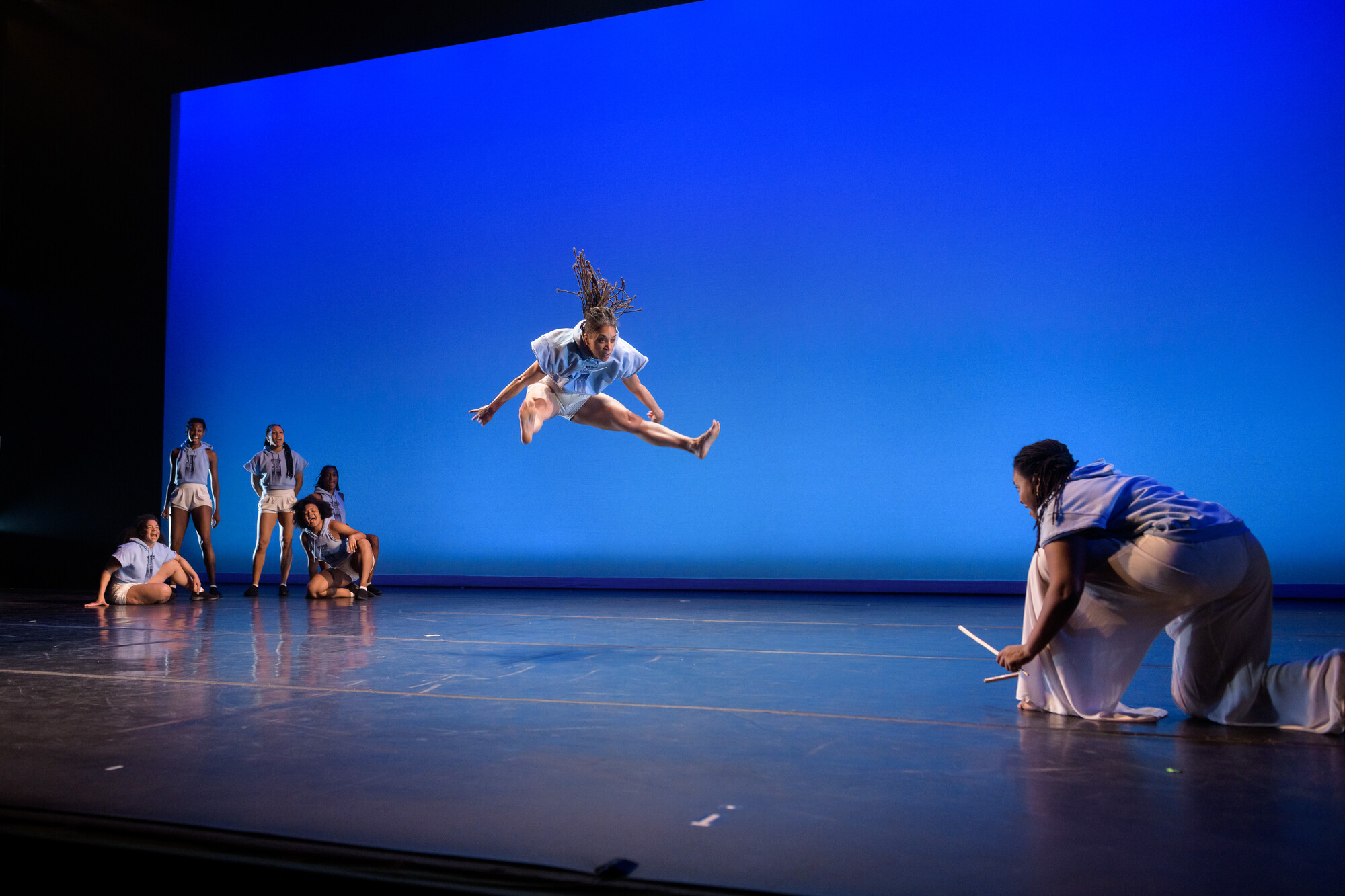Four performers sit around a dancer jumping in front of a blue background.