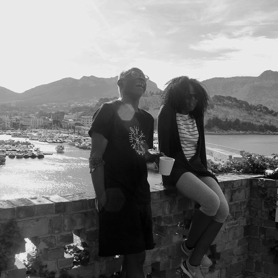 A man and a woman lean against a stone wall overlooking a body of water and mountains.