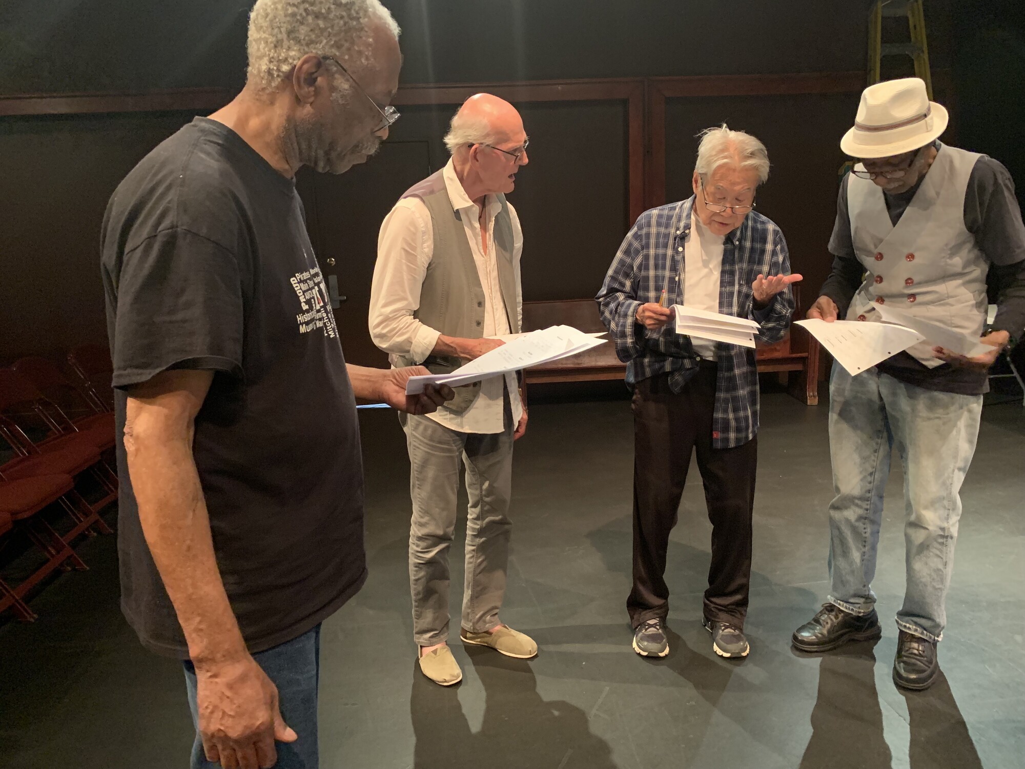 Four senior actors stand on-stage and look at their scripts during a rehearsal.