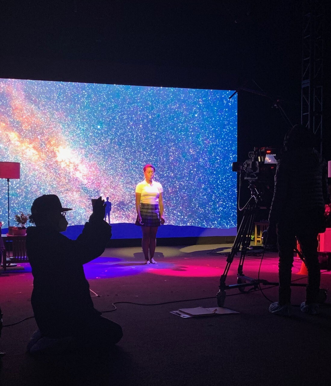 A performer stands in front of a projection screen while crew members set up cameras and lights.