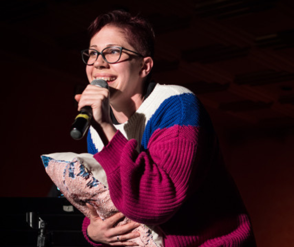 A woman wearing glasses and a red sweater holding a pillow and speaking into a microphone.