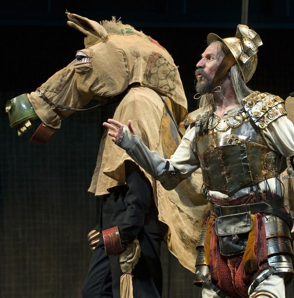 A man in full armor stands next to a puppet horse on stage.