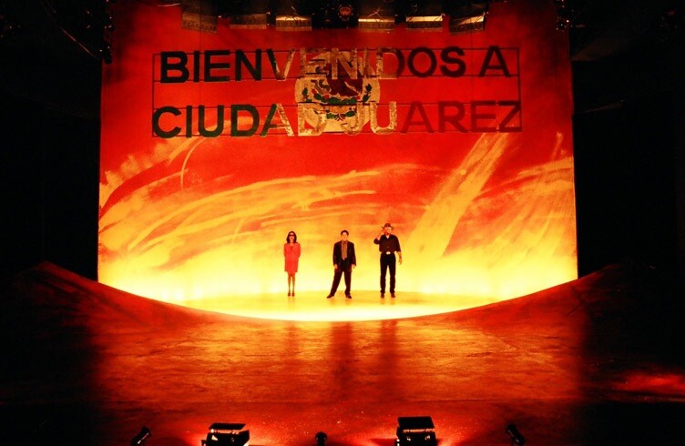 Three performers stand in front of a fire-like backdrop where a sign above them reads "Bienvenidos a Ciudad Juarez."