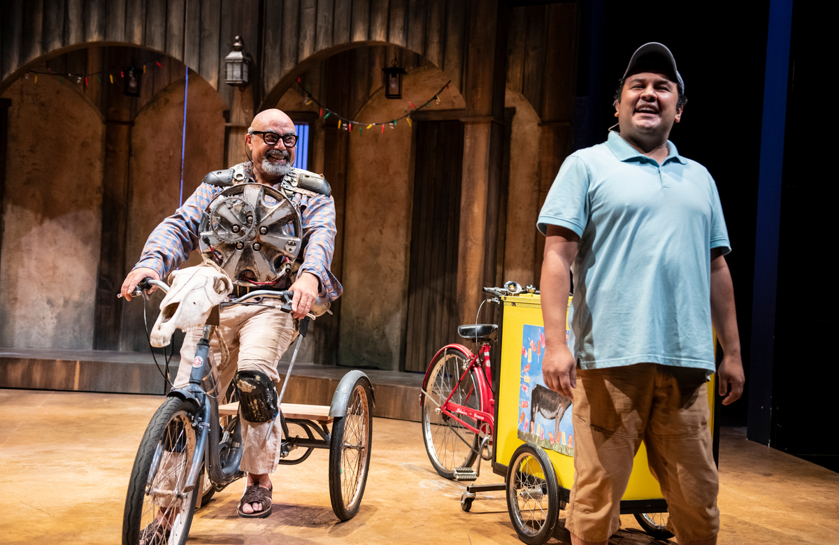 A man on a tricycle smiles on stage with another actor.