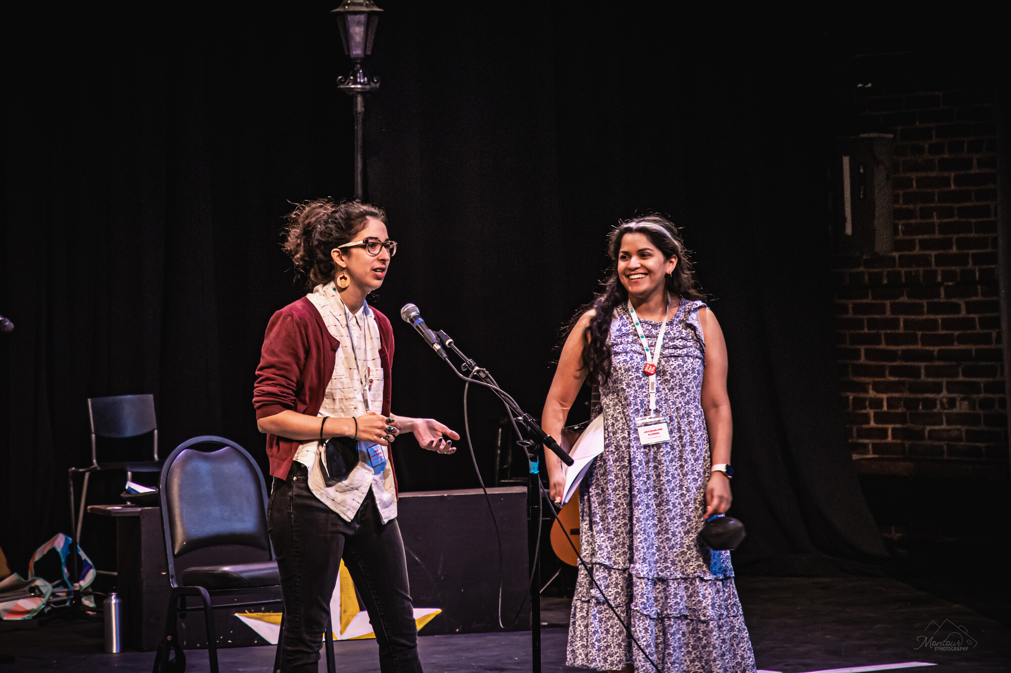 One woman speaks into a microphone and another woman stands next to her and smiles.