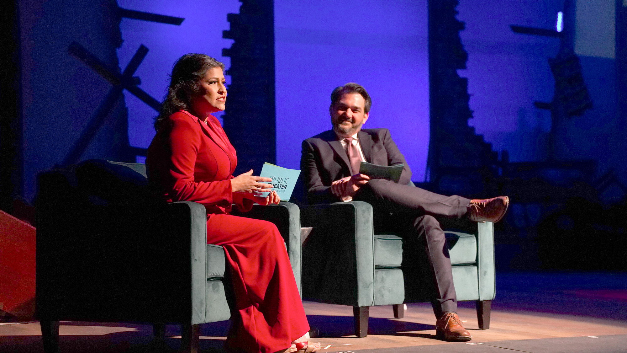A woman and a man sit in armchairs and talk on a lit stage.