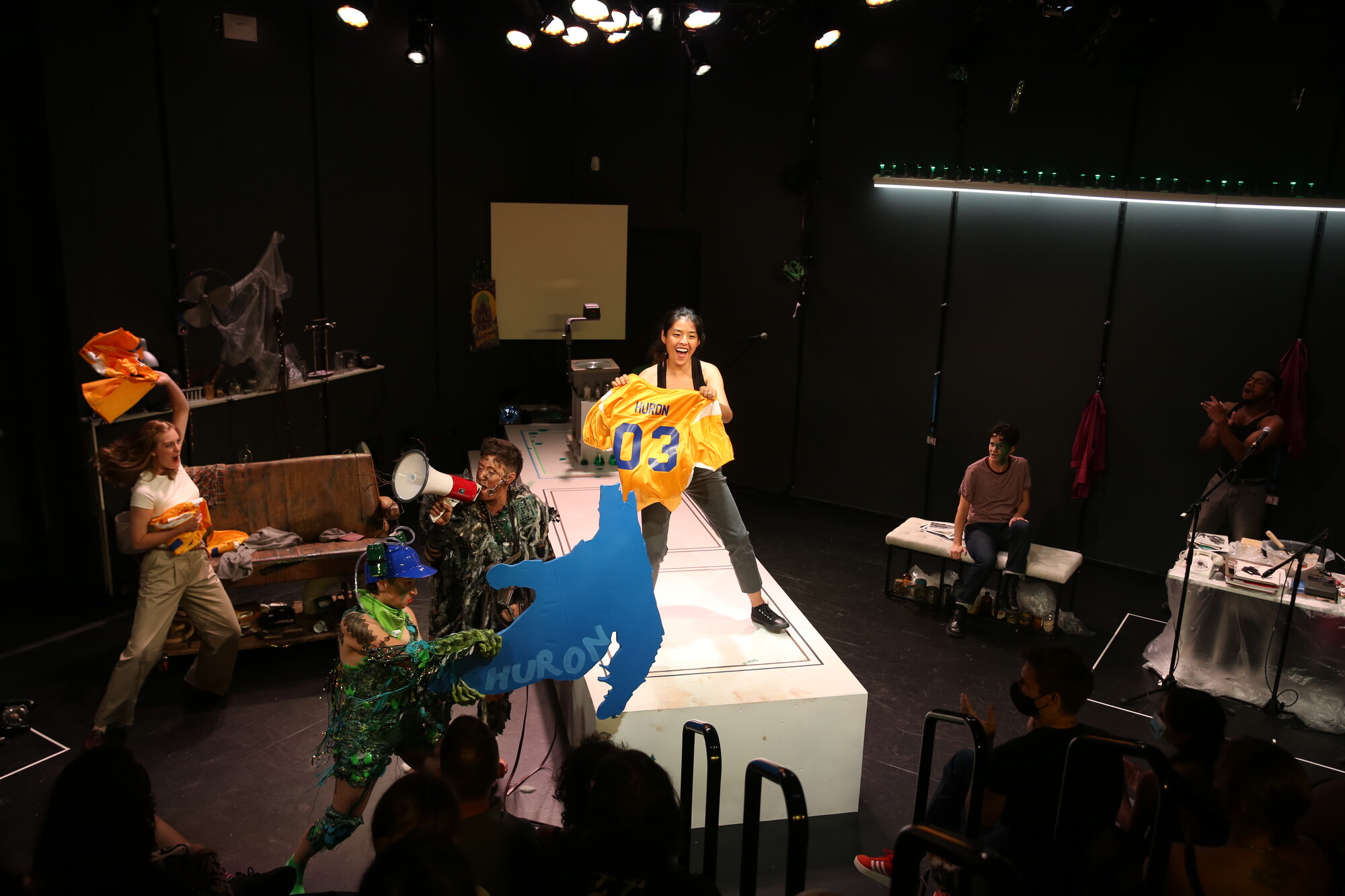 A performer holds up a prop version of Lake Huron while another dances with a sports jersey with "Huron" on the back and a third yells into a megaphone.