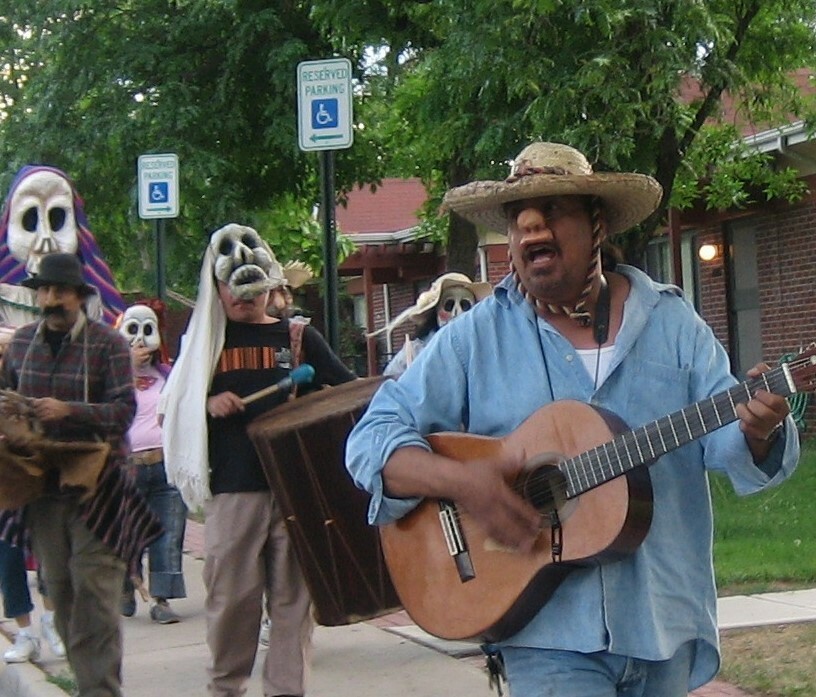 A man plays the guitar while walking down the street and wearing a pig mask with several other costumed people behind him.
