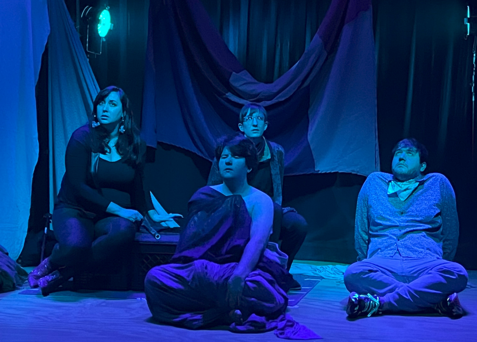 Four people sit under a blue light, one closes their eyes and three look up to the sky.
