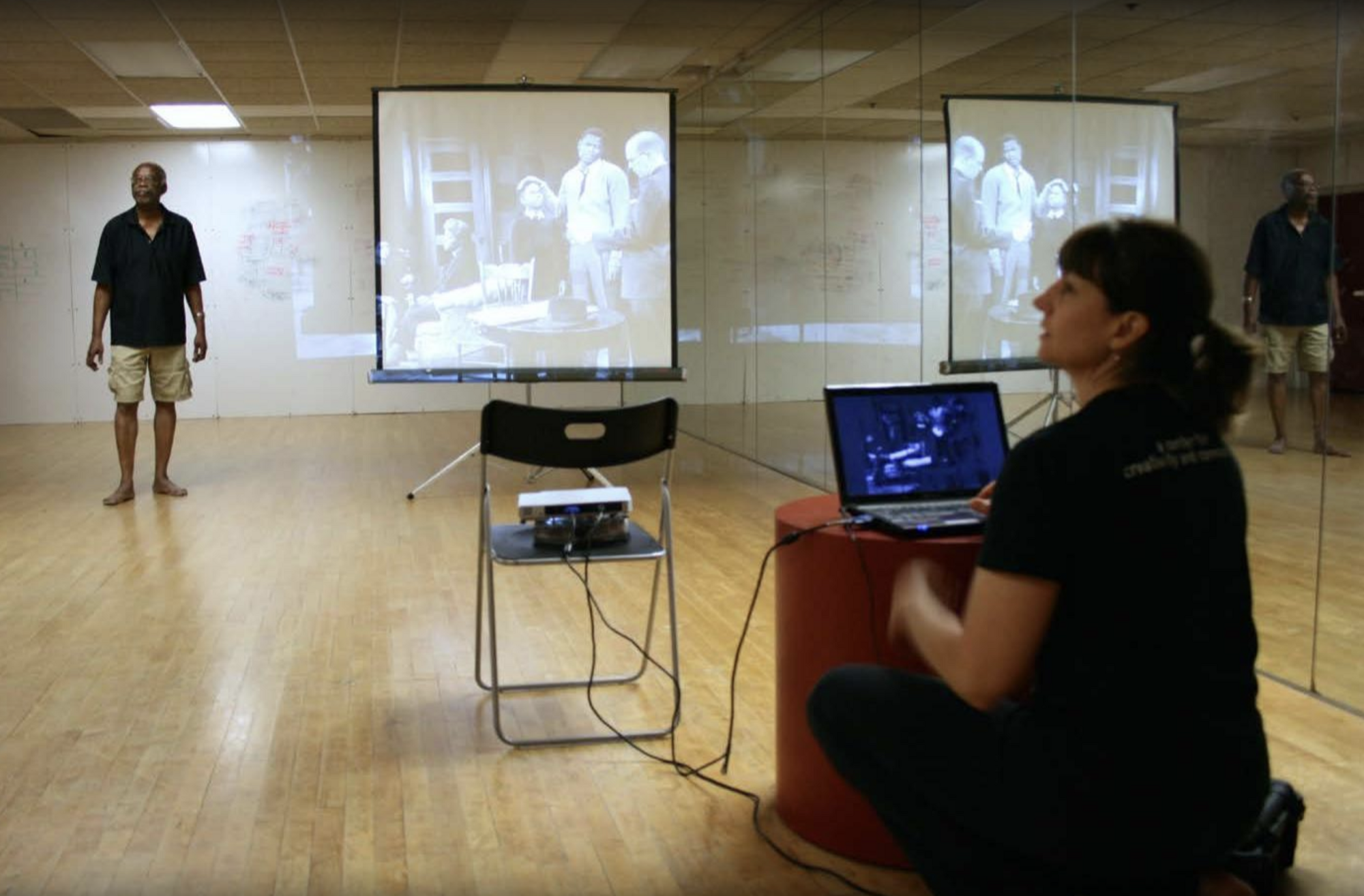 In a mirrored rehearsal room, a man stands next to a projection screen while a woman sits with a laptop and manages the images being shown on the projector.