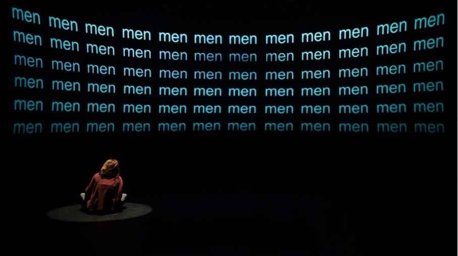A man sits on stage in the dark, looking up at a projection of a grid that reads "men" over and over again.