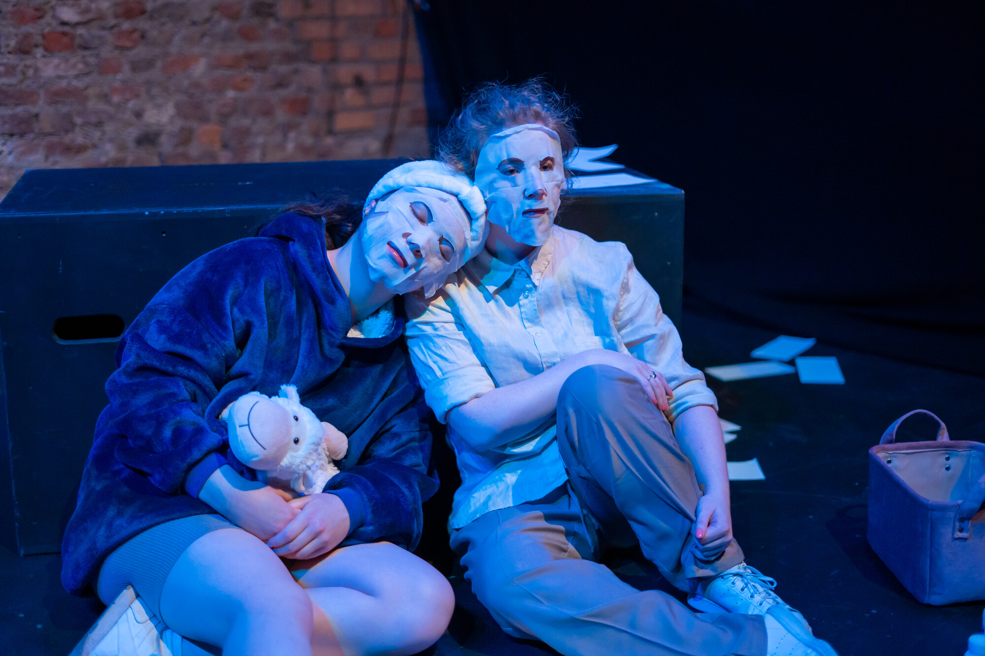 Two actors wearing face masks sit and cuddle together on stage.