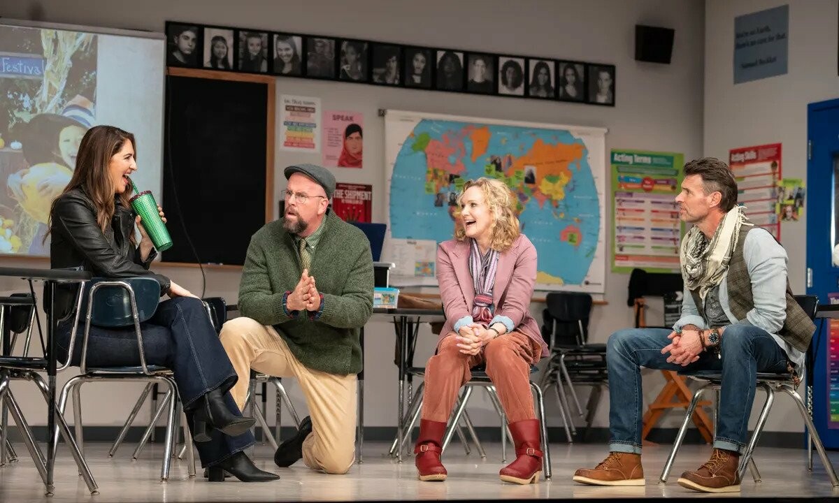 Four actors sit on chairs on a set made to look like a children's classroom, smiling and talking with one another.