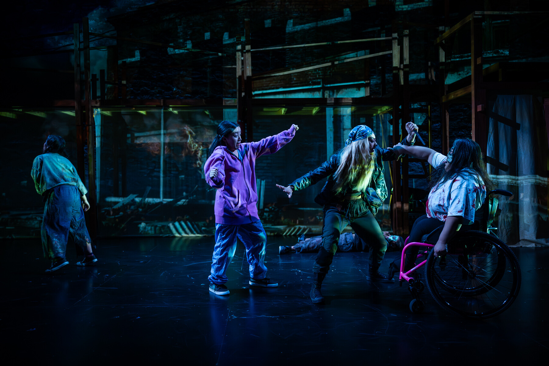 On stage, a performer in a wheelchair grabs the arm of another performer in a stage fight, while three other performers in various states of shock or injury are scattered around the space.