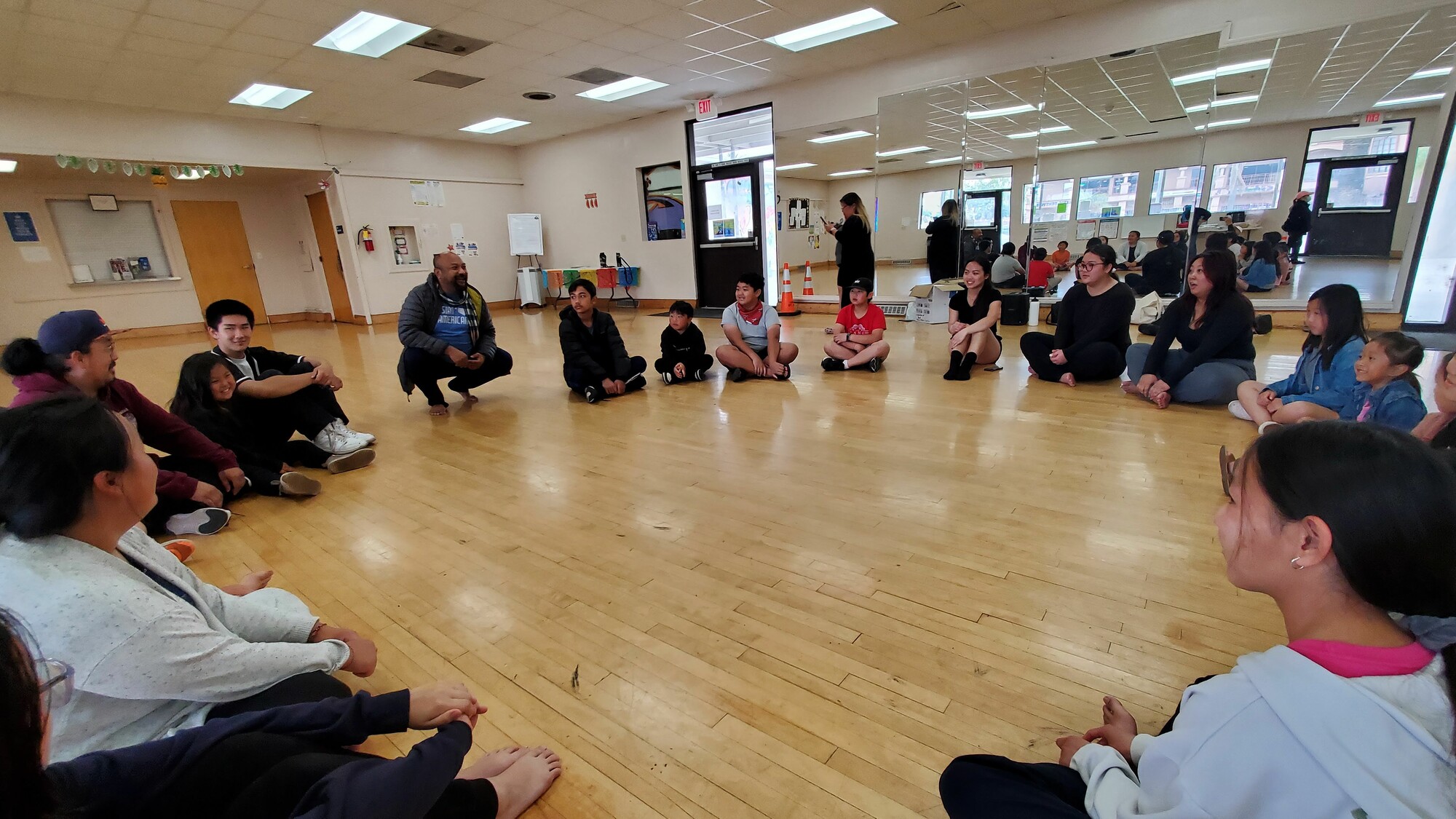 In a large mirrored rehearsal room, two adult facilitators and numerous young people sit in a circle and smile. 